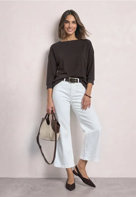 Frau mit dunkelbraunem Shirt, weißen Culottes, flachen Schuhen und Tasche vor hellem Hintergrund.