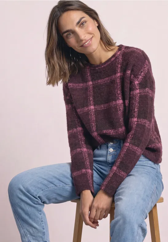 Frau auf Hocker sitzt, trägt karrierten bordeauxroten Pullover und blaue Jeans, lächelt leicht.