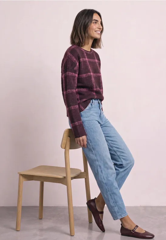 Eine junge Frau in lila Pullover und Jeans lehnt entspannt an einem Stuhl vor rosa Hintergrund.