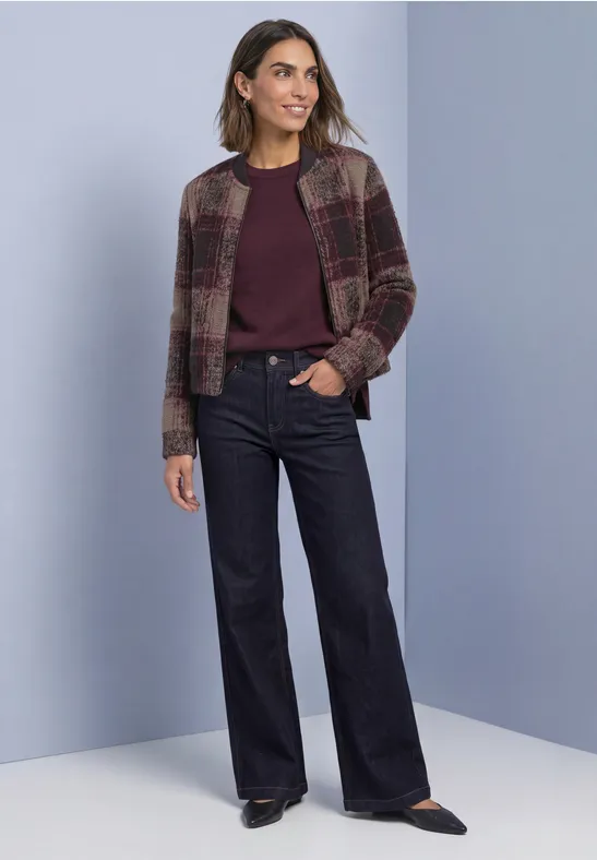 Vrouw voor lichte achtergrond, paarse trui, geruite jas en wijde donkere jeans.