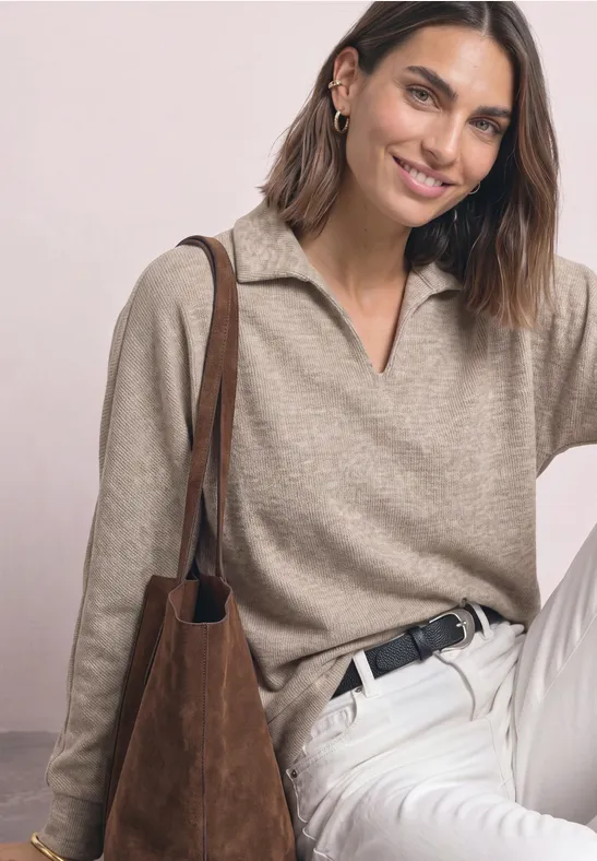 Lächelnde Frau in beigefarbenem Pullover, weißen Hosen und brauner Tasche vor hellem Hintergrund