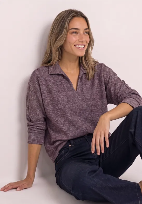 Eine junge Frau in lila Pullover und dunkler Jeans sitzt entspannt auf dem Boden vor einer hellen Wand.