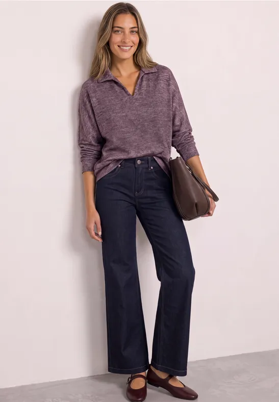 Frau in lila Pullover und dunkelblauen Jeans mit brauner Tasche vor weißer Wand.