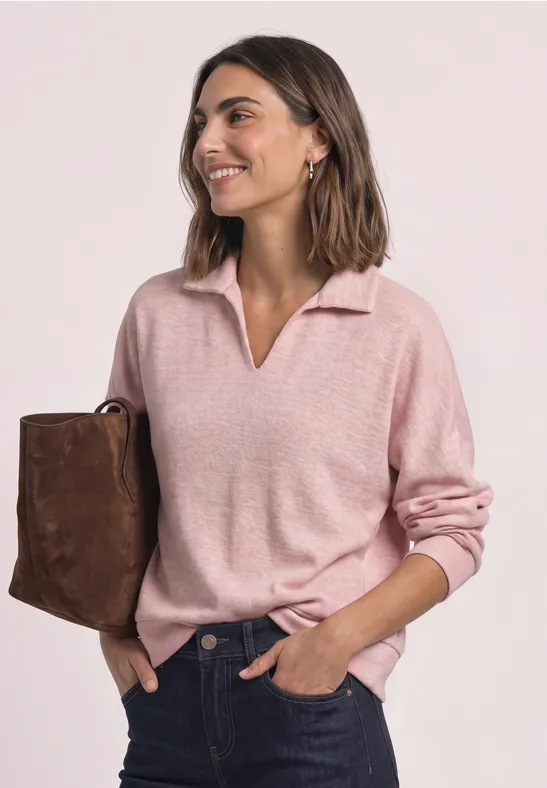Frau in rosa Pullover mit V-Ausschnitt und brauner Umhängetasche lächelt seitlich mit Händen in den Taschen