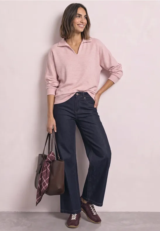 Frau trägt rosa Pullover und weite Jeans, hält eine braune Ledertasche mit kariertem Schal.