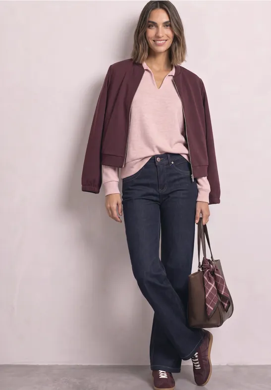Frau in lässig-elegantem Outfit: bordeauxfarbene Jacke, rosafarbener Pullover, dunkelblaue Jeans, Tasche und Schuhe in Braun.