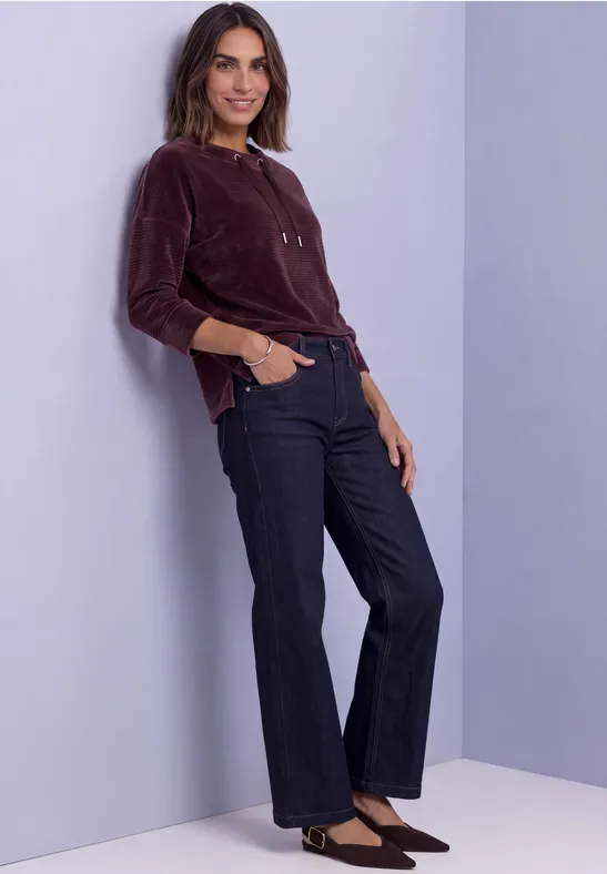 Vrouw leunt glimlachend tegen een lila muur, draagt een bordeaux top, donkere jeans en zwarte pumps.