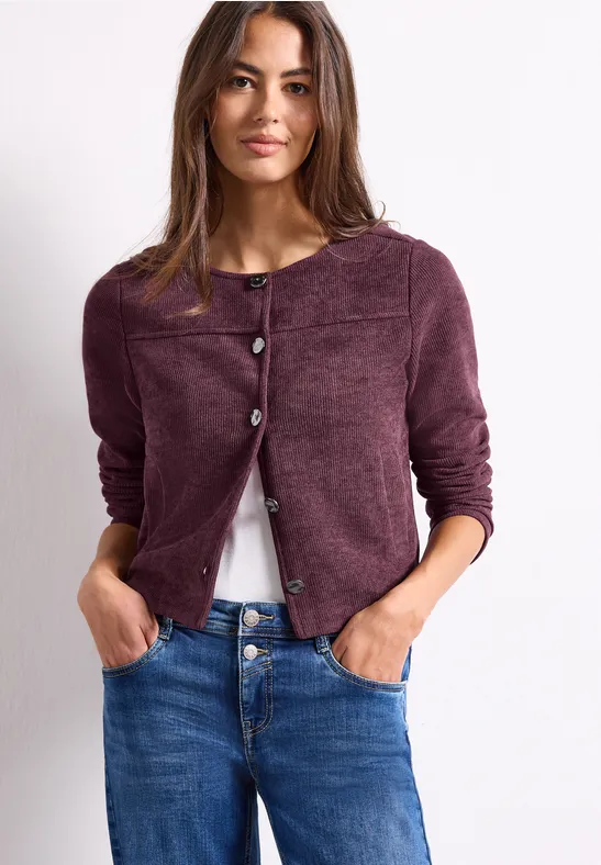 Frau in lila Strickjacke, weißem Shirt und Jeans, Hände in Taschen, lässiger Stil