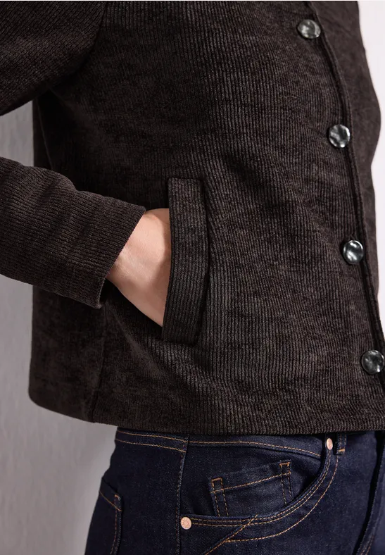 Dunkelbraune Jacke mit Knöpfen, Hand in der seitlichen Tasche, dazu blaue Jeans.