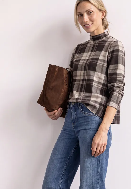 Eine Frau in karierter Bluse und Jeans hält eine braune Tasche unter dem Arm vor hellem Hintergrund.