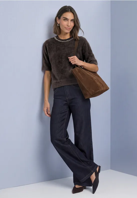 Frau in braunem Kurzarmshirt, weitem Jeansbund, schwarzen Ballerinas und brauner Tasche vor hellblauem Hintergrund