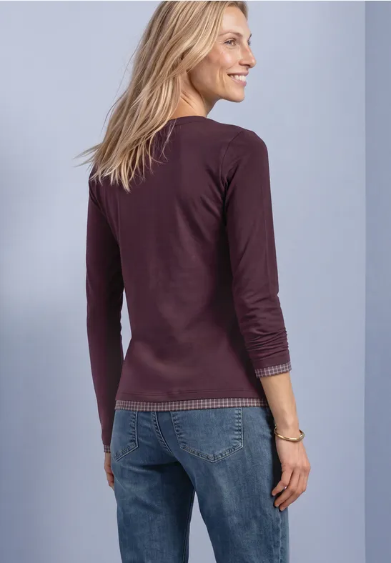 Eine lächelnde Frau von hinten, trägt lila Langarmshirt und blaue Jeans vor hellem Hintergrund.