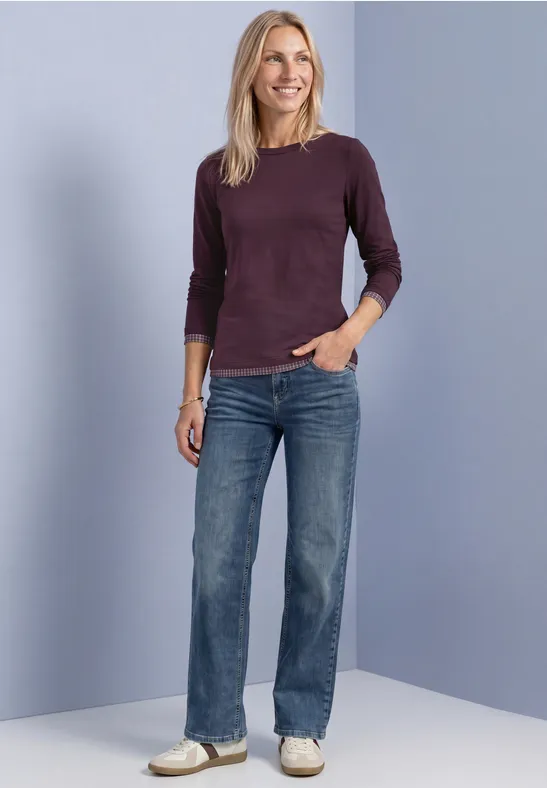 Eine Frau in Jeans, beerenfarbenem Pullover und weißen Sneakern vor hellem, bläulichem Hintergrund.