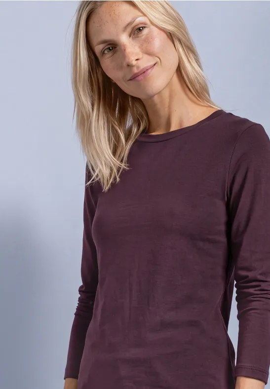 Eine lächelnde blonde Frau in plumafarbenem Langarmshirt vor helllila Hintergrund.