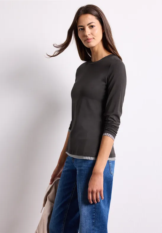 Eine Frau in schwarzem Langarmshirt und Jeans hält eine Jacke vor weißem Hintergrund.