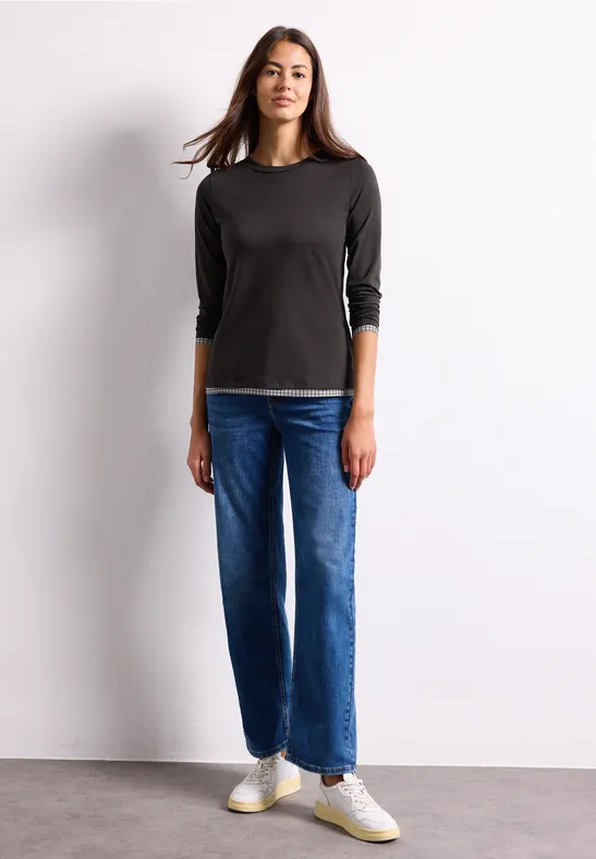 Junge Frau steht vor weißer Wand, trägt schwarzen 3/4-Ärmel-Pullover, blaue Jeans und weiße Sneaker.