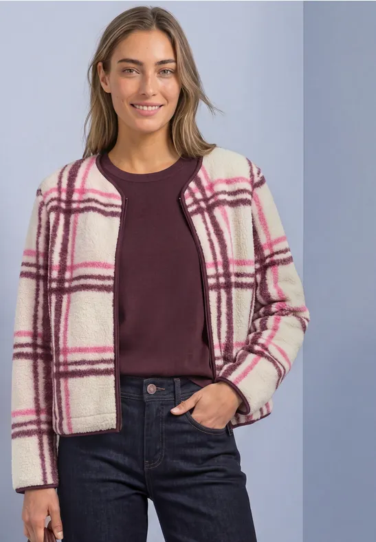 Vrouw draagt witte jas met roze ruitpatroon, bordeaux top en donkere jeans voor lichte achtergrond.