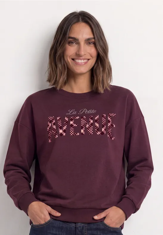 Frau in bordeauxrotem Pullover mit Blütenstickerei und Schriftzug.