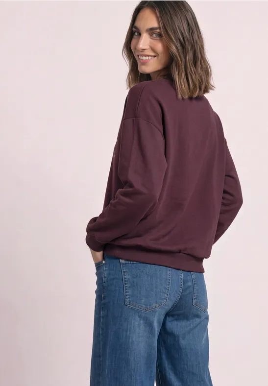 Frau mit weinrotem Pullover und blauen Jeans, lächelnd über die Schulter blickend.