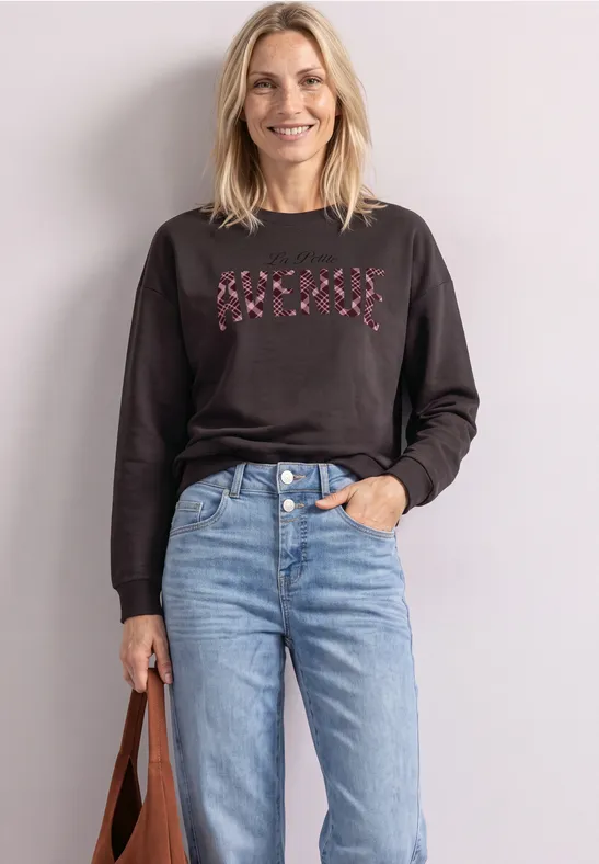 Eine lächelnde Frau trägt schwarzen Pullover mit rosa Kreuzmotiv, hellblaue Jeans und hält eine braune Tasche.
