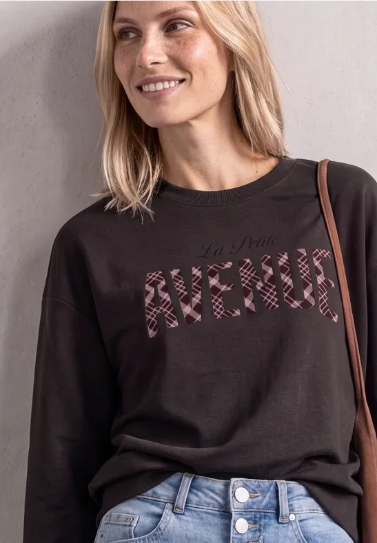 Eine lächelnde Frau trägt einen schwarzen Pullover mit zebrastreifenem Schriftzug "AVENUE" und blaue Jeans.