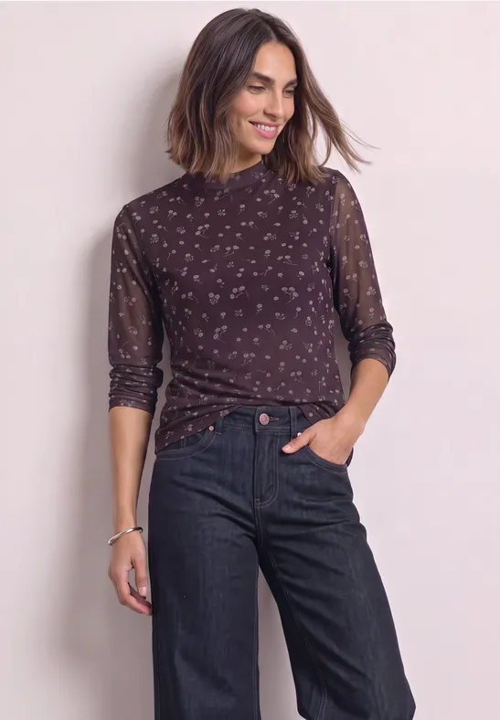 Frau mit halblangem dunklem, gemustertem Oberteil und dunklen Jeans, die lächelnd in die Ferne schaut.