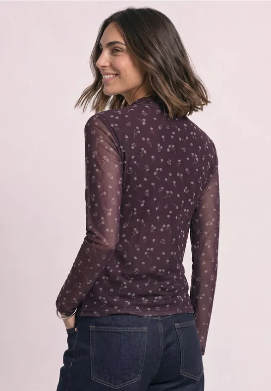 Frau in Jeans und transparentem dunkelrotem Langarmshirt mit kleinem Blumenmuster, lächelnd seitlich stehend.