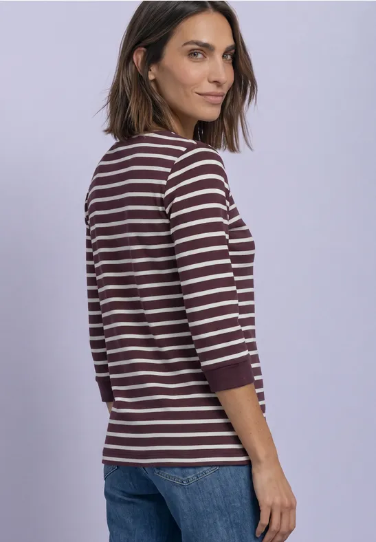 Eine Frau in lila-weiß gestreiftem Langarmshirt und Jeans vor hellem violettem Hintergrund.