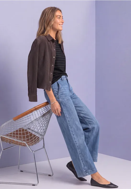 Frau in brauner Jacke, schwarzem Shirt und weiten Jeans lehnt lässig an einem Drahtstuhl vor lila Wand.