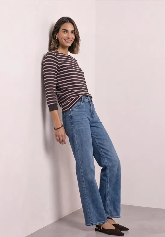 Eine Frau in gestreiftem Langarmshirt und Jeans lehnt lächelnd an einer Wand.