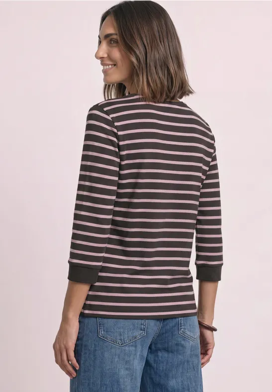 Frau mit braunen Haaren von hinten, trägt gestreiftes Langarmshirt und Jeans, im Profil vor rosa Hintergrund.