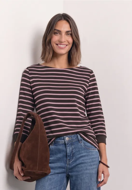 Lächelnde Frau trägt gestreiftes Langarmshirt, blaue Jeans und braune Wildleder-Umhängetasche.