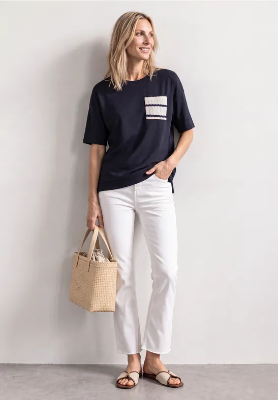 Vrouw in casual outfit: donker T-shirt met gestreepte zak, witte broek, sandalen en een strooien tas.