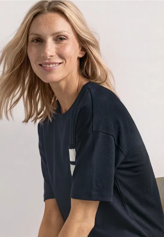 Blonde vrouw lacht in een donker T-shirt, zittend schuin en kijkend naar de camera.