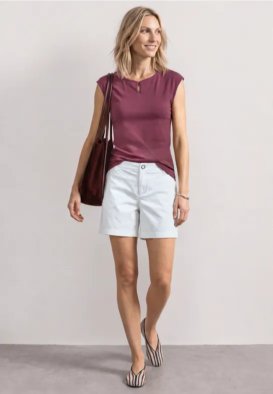 Vrouw met bordeaux top, witte short en slippers, draagt grote tas voor neutrale achtergrond.
