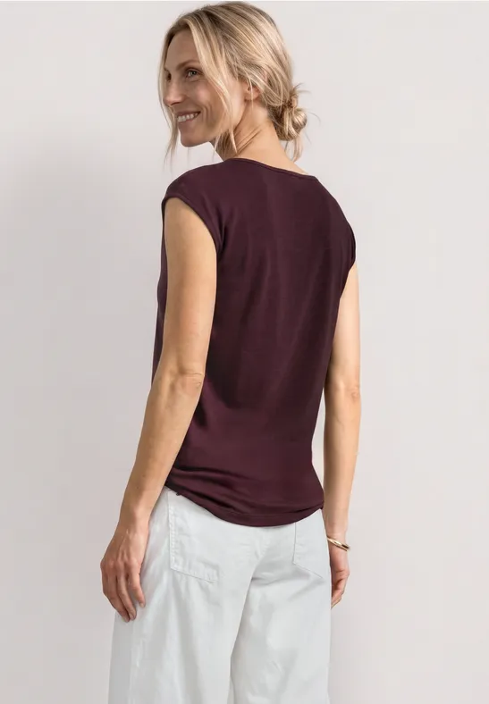 Vrouw van achteren in een bordeauxrood mouwloos shirt en losse witte broek tegen lichte achtergrond.