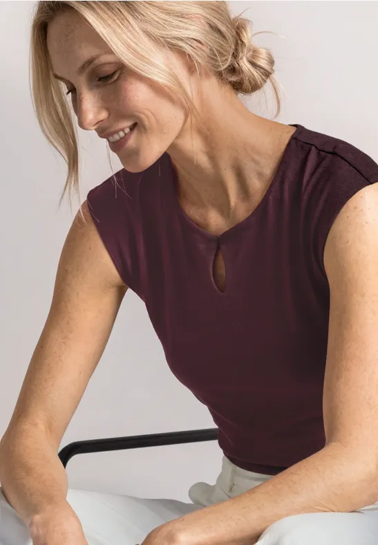 Een zittende vrouw met opgestoken blond haar en een donkerrode mouwloze top.