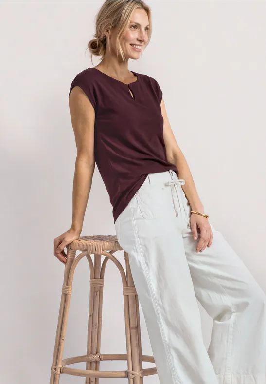 Vrouw leunt op een houten kruk, draagt een bordeaux kort shirt en een lichte, wijde broek.