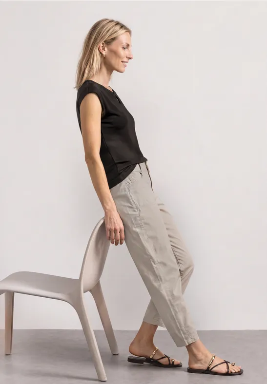 Vrouw in zwart shirt en beige broek leunt zijwaarts tegen een stoel, draagt platte sandalen.