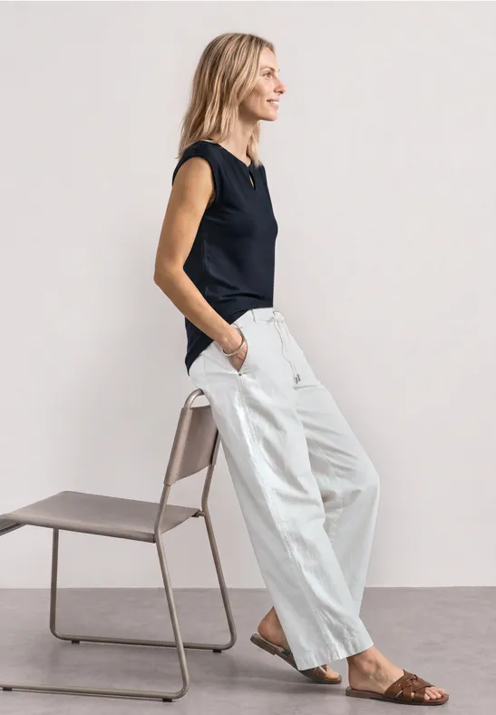 Une femme en haut noir, pantalon blanc ample et sandales pose de profil près d'une chaise minimaliste.