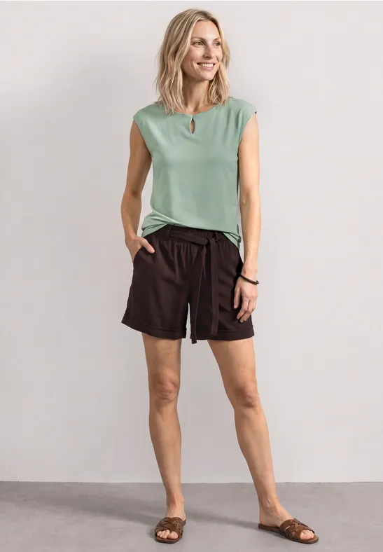 Une femme en haut vert menthe sans manches, short noir et sandales brunes pose devant un mur neutre.