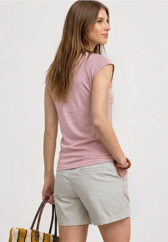 Femme de dos en t-shirt rose et short beige, tenant un sac à main marron devant un mur clair.