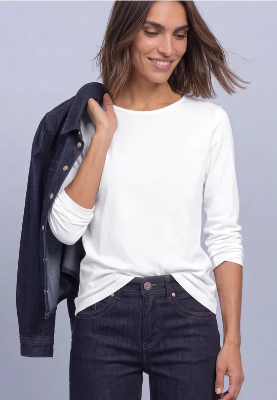 Vrouw in wit longsleeve-shirt met donker spijkerjasje op de schouder en donkere spijkerbroek, glimlachend.