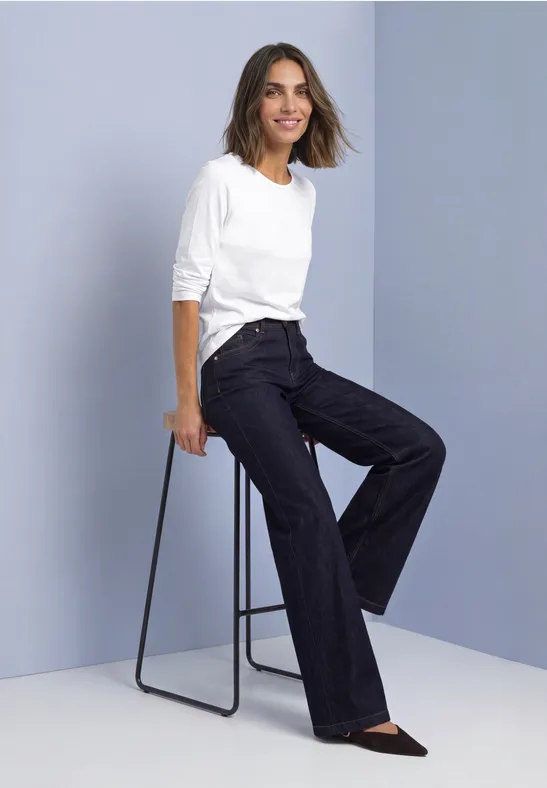Vrouw zijdelings zittend op krukje, witte blouse, losse donkere broek en zwarte schoenen.