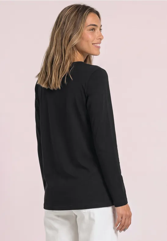 Frau in schwarzem Langarmshirt und weißer Hose, steht seitlich vor hellem, rosafarbenem Hintergrund.