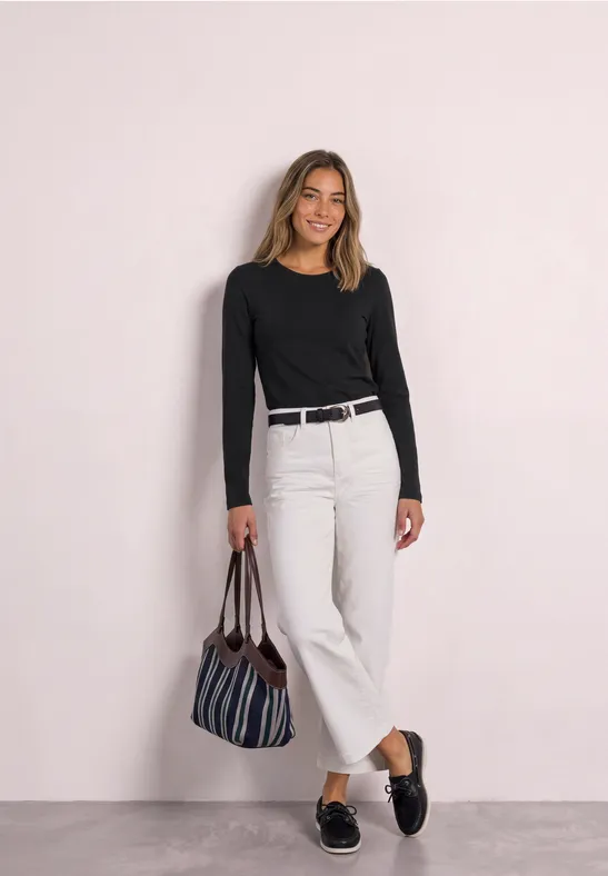 Frau in schwarzem Langarmshirt, weißer weiter Hose, schwarzem Gürtel, gestreifter Tasche und Loafers lächelnd.