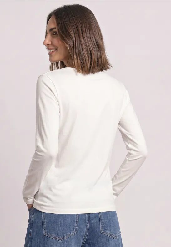 Frau von hinten in weißem Langarmshirt und blauen Jeans vor neutralem Hintergrund.
