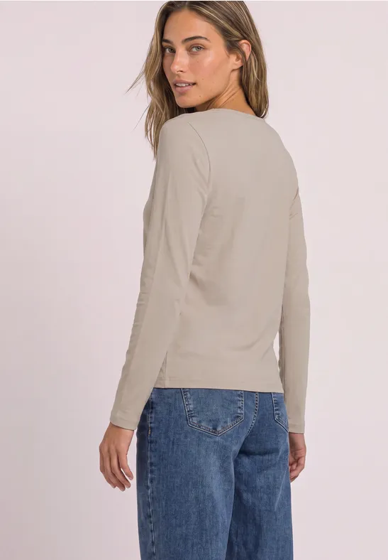 Frau trägt beiges Langarmshirt und blaue Jeans, schaut über die Schulter vor hellem Hintergrund.