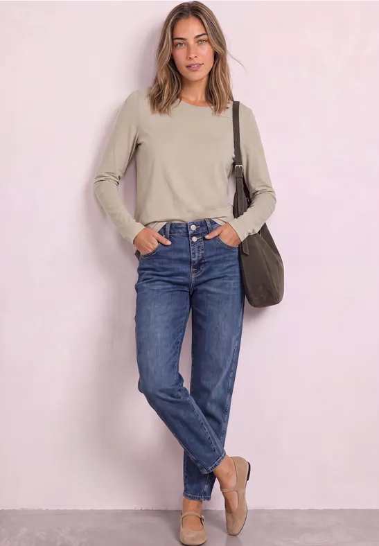 Junge Frau in beige Longsleeve, blauen Jeans und Loafers lehnt entspannt vor hellrosa Wand.