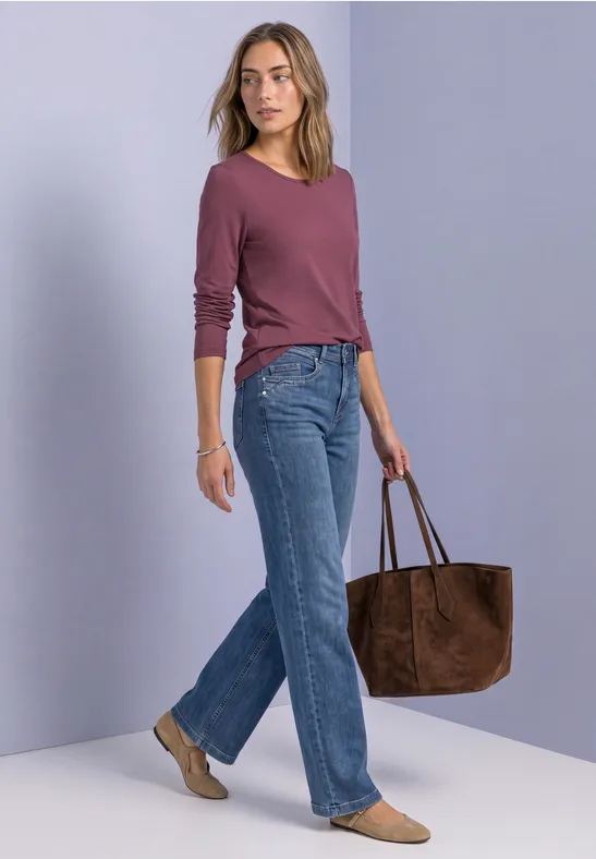 Frau in lila Oberteil, blauen Jeans und beigen Schuhen trägt eine große braune Wildledertasche.
